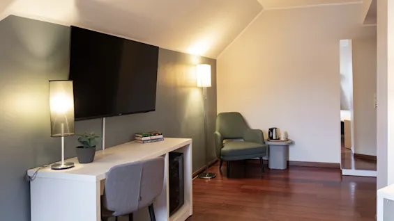 Ett mysigt rum med ett skrivbord och TV på ena sidan, och en bekväm fåtölj med en lampa på den andra, perfekt för arbete eller avkoppling.