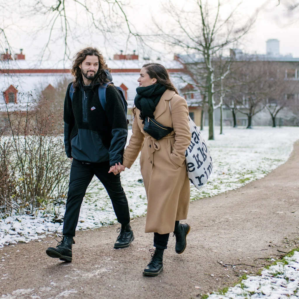 Couple walking hand-in-hand in snowy Kaivopuisto in Helsinki, with buildings in the background. The woman carries a bag with text.