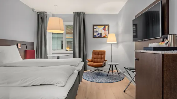 En pænt redt seng i et moderne hotelværelse vender mod et vægmonteret TV. Nærved står en lænestol og en lampe på et rundt tæppe. Et vindue og kunstværker pryder væggene.