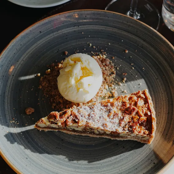 En bit nötpaj serverad på en grå tallrik med en kula vaniljglass över smuldeg.