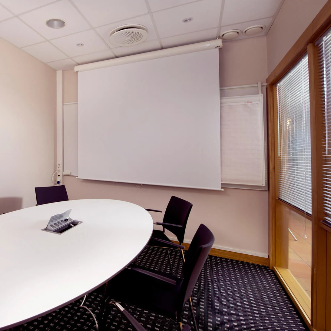 Et konferanserom med et ovalt bord, stoler og et hvitt projeksjonslerret i et rent, opplyst rom.