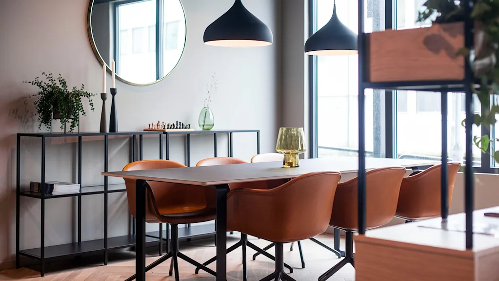 Spisebord omgitt av brune stoler, plassert i et moderne rom med store vinduer. Over henger to svarte pendellamper, mens planter og dekorasjoner pryder hyller mot veggen.