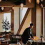 En person arrangerer et bord i en varmt oplyst, moderne restaurant med hængelamper og store vinduer. Bordene er dækket med glas og omgivet af hyggelige siddepladser.