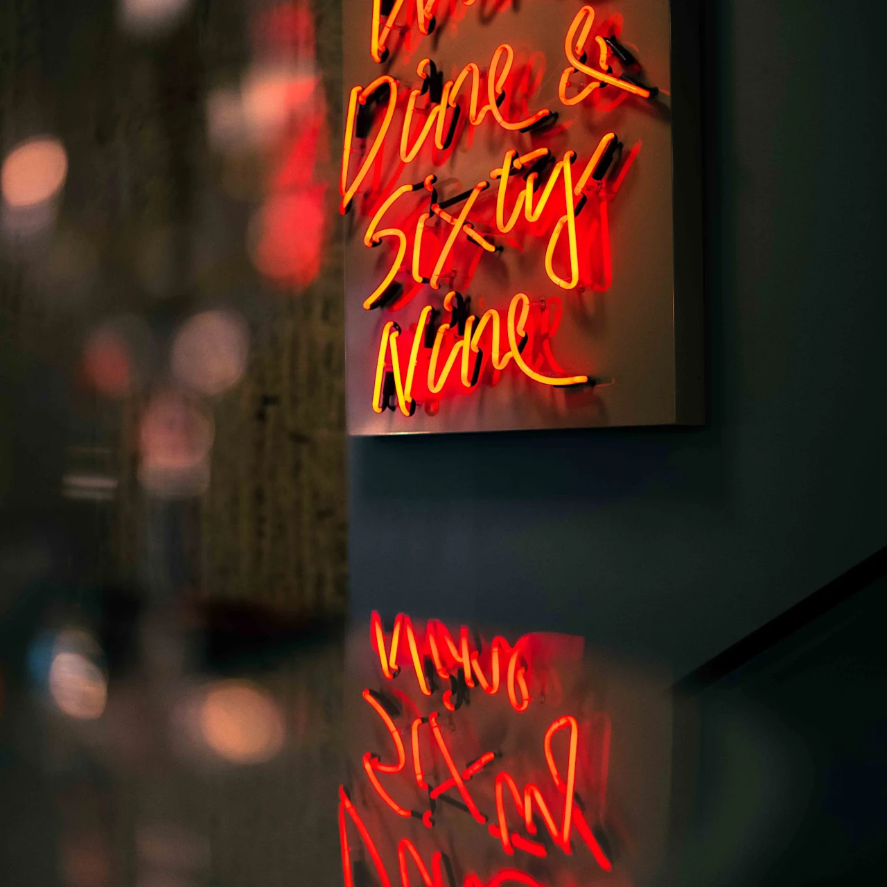 A neon sign glows with red cursive letters reading "Wine Dine & Sixty Nine," reflecting on a nearby glossy surface in a dimly lit setting.