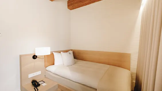 Ett bekvämt och minimalistiskt hotellrum med en enkelsäng, ett litet nattduksbord och takdetaljer i trä.