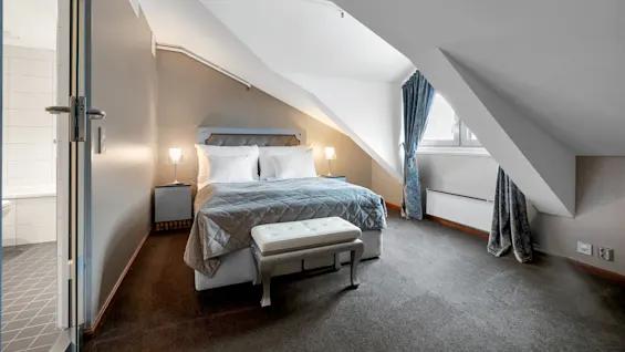 En seng med hvide puder og et gråt tæppe står under et skrånende loft i et hyggeligt soveværelse. Blå gardiner dækker et vindue ved siden af, med en bænk for enden.