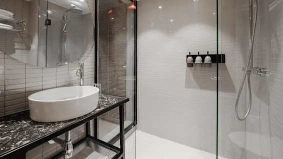 A modern bathroom features a round sink on a black counter, a circular mirror, and a glass shower enclosure. White tiled walls and overhead lighting create a bright atmosphere.