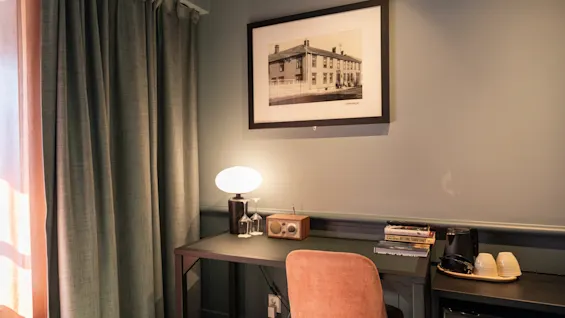 Et skrivebord med en lampe, radio og bøger står i et rum ved siden af trukne gardiner. Et indrammet foto af en bygning hænger på væggen ovenfor.