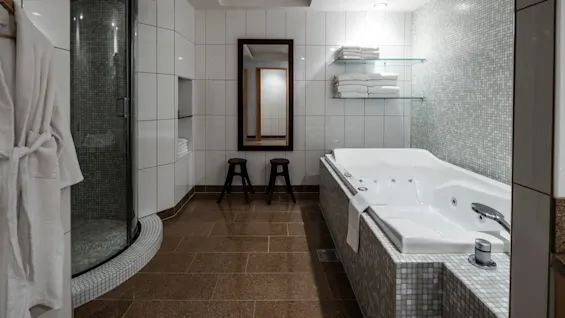 Lyxigt vitt badkar står stilla i ett rymligt badrum med kaklade väggar, glasdusch och staplade handdukar på hyllor. Två pallar och en spegel förstärker den moderna atmosfären.