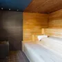 Træbænke kant linjerer en spasauna med et håndklæde og lotion placeret på den øvre bænk; mørke strukturerede vægge og en stenvarmer forbedrer den rolige atmosfære.