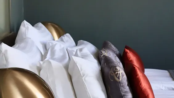 A bed with neatly arranged pillows in white, purple, and red, accompanied by a brass lamp. The scene is set against a gray wall adorned with framed photographs.