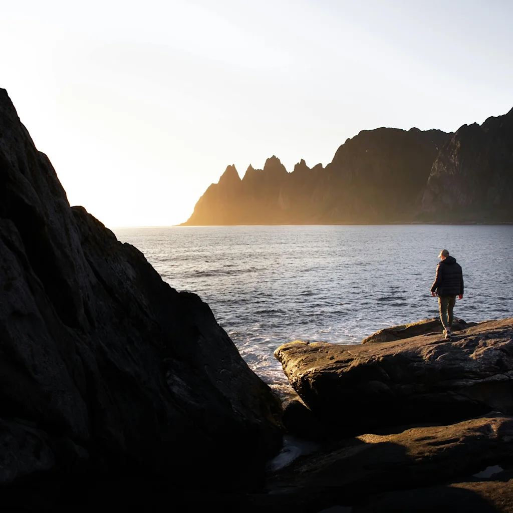 En person står på en klippefyldt kyst og ser ud over havet og fjerne bjerge under en lys solnedgang.