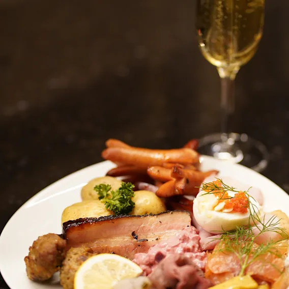 En velsmagende tallerken med blandede kødtyper, æg og garneringer er placeret på et bord, ledsaget af et højt glas mousserende vin i en svagt oplyst restaurantindstilling.