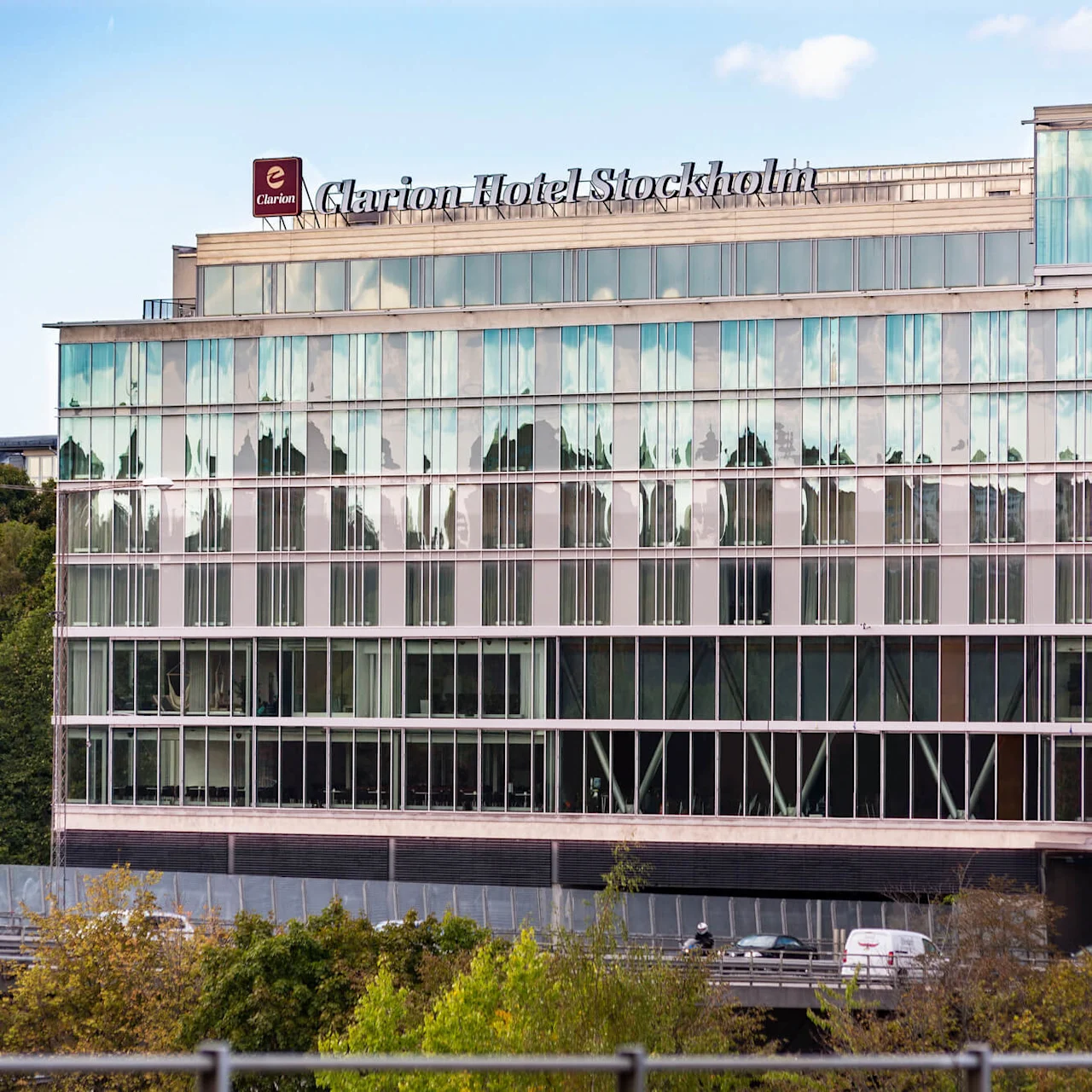 En modern hotellbyggnad visar skylten "Clarion Hotel Stockholm" ovanför reflekterande glasfönster. Träd och en väg med en bil finns i förgrunden.