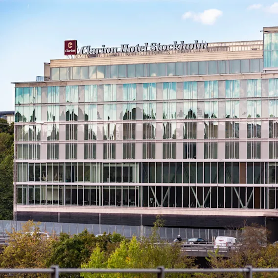 A modern hotel building displays the sign "Clarion Hotel Stockholm" atop reflective glass windows. Trees and a road with a car appear in the foreground.