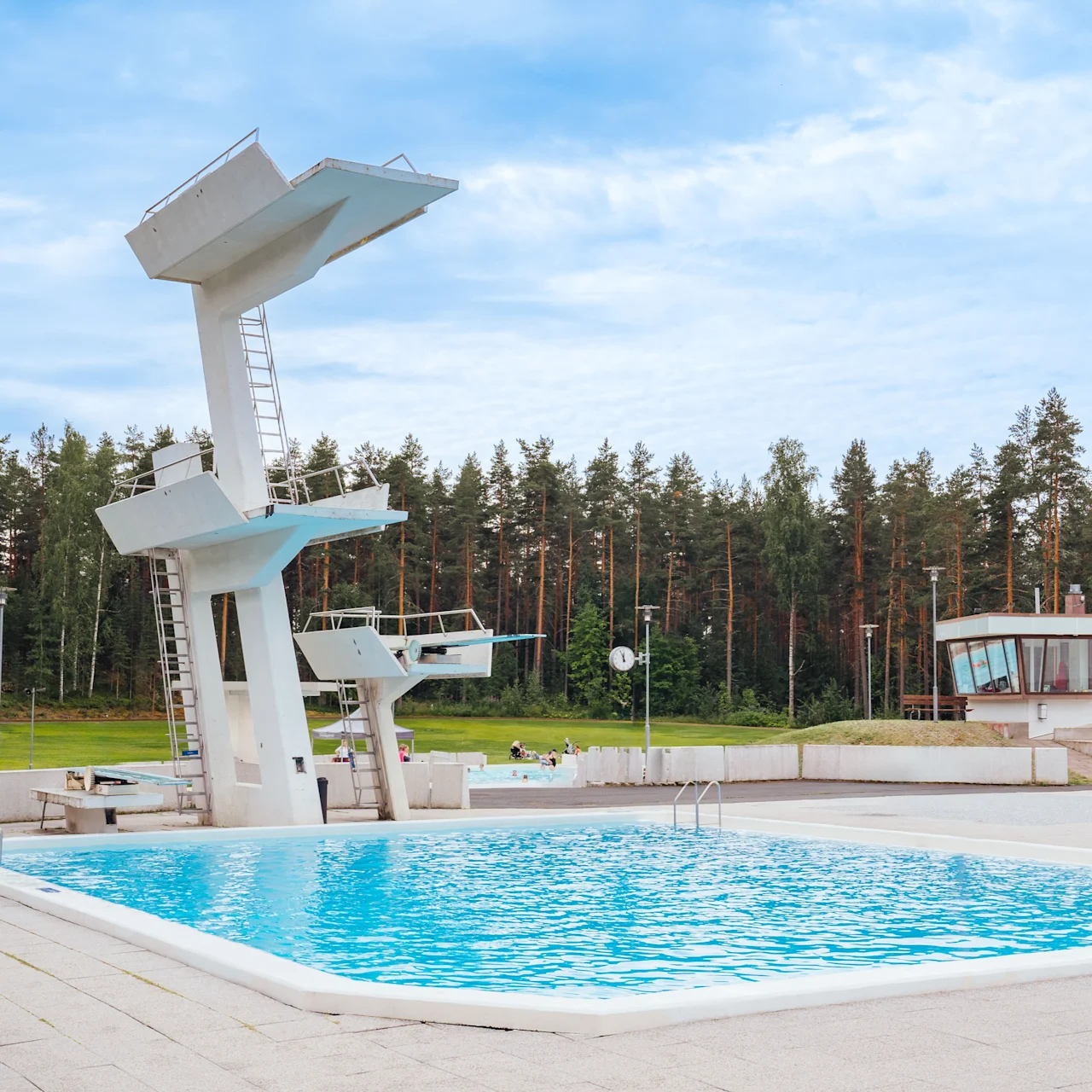 A large outdoor swimming pool with multiple diving platforms, surrounded by a forest of tall trees under a cloudy sky. A small building is visible in the background.