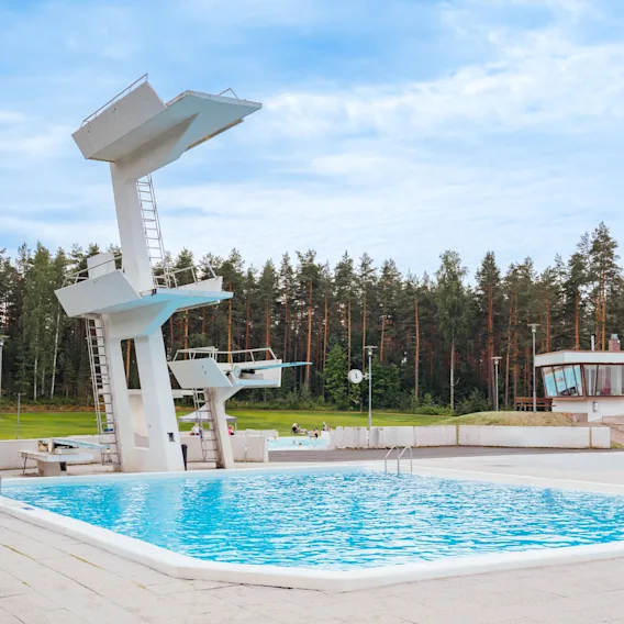 Suuri ulkouima-allas, jossa on useita uimahyppyalustoja, ympäröity korkealla metsällä pilvisen taivaan alla. Taustalla näkyy pieni rakennus.