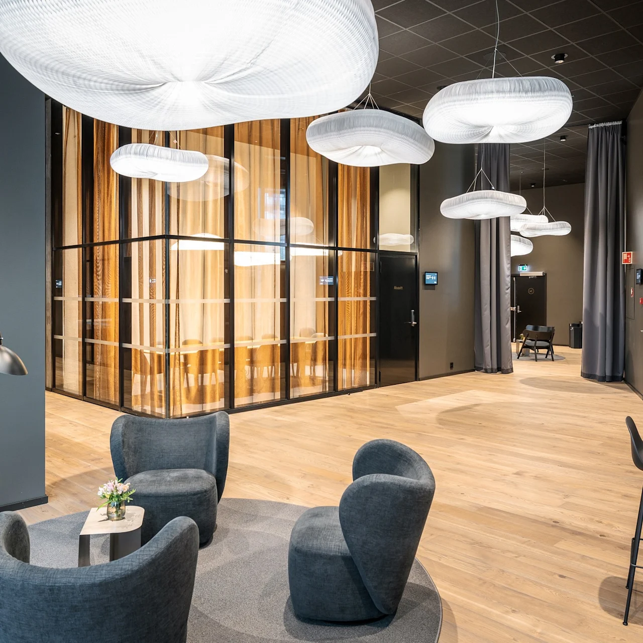 Bekväma grå stolar omger ett litet bord med blommor i en modern lounge. Upphängda vita lampor hänger ovanför och förstärker trägolvet och glasväggarna i mötesrummet.