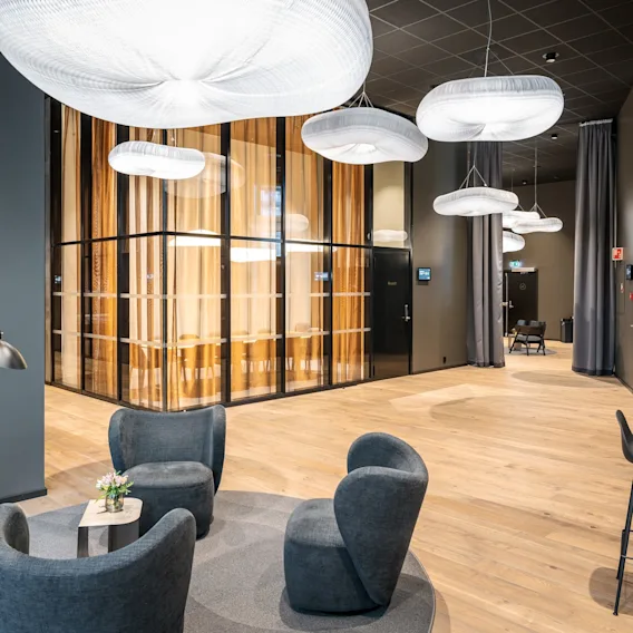 Komfortable grå stoler omgir et lite bord med blomster i en moderne lounge. Hengende hvite lys henger over, og fremhever tregulvet og glassveggene i møterommet.