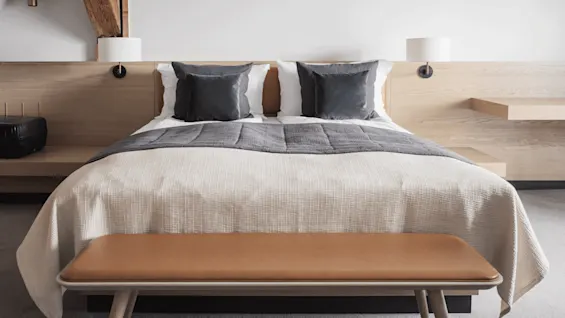 A neatly made bed with gray and white bedding sits between two tables, under a sloped wooden beam, in a modern, minimalist bedroom.