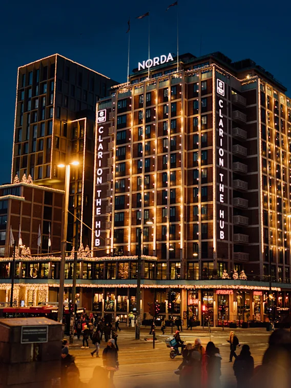 En fleretasjes bygning, opplyst av lys, viser tekstene "CLARION THE HUB" og "NORDA" om natten. Den er omgitt av mennesker som går i et travelt byområde.