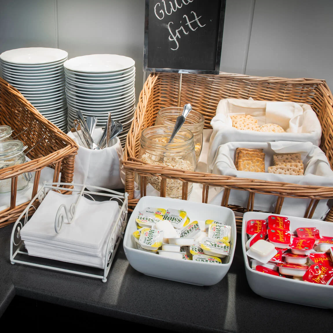 Frukostobjekt på buffé presenteras inklusive flingor i burkar, högar av tallrikar och förpackningar av smör och sylt, med en skylt som läser "Glutenfritt".