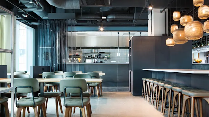 En moderne kaféinteriør med trebord og -stoler, hengende lamper og åpent kjøkken, formidler en stilig og innbydende atmosfære.