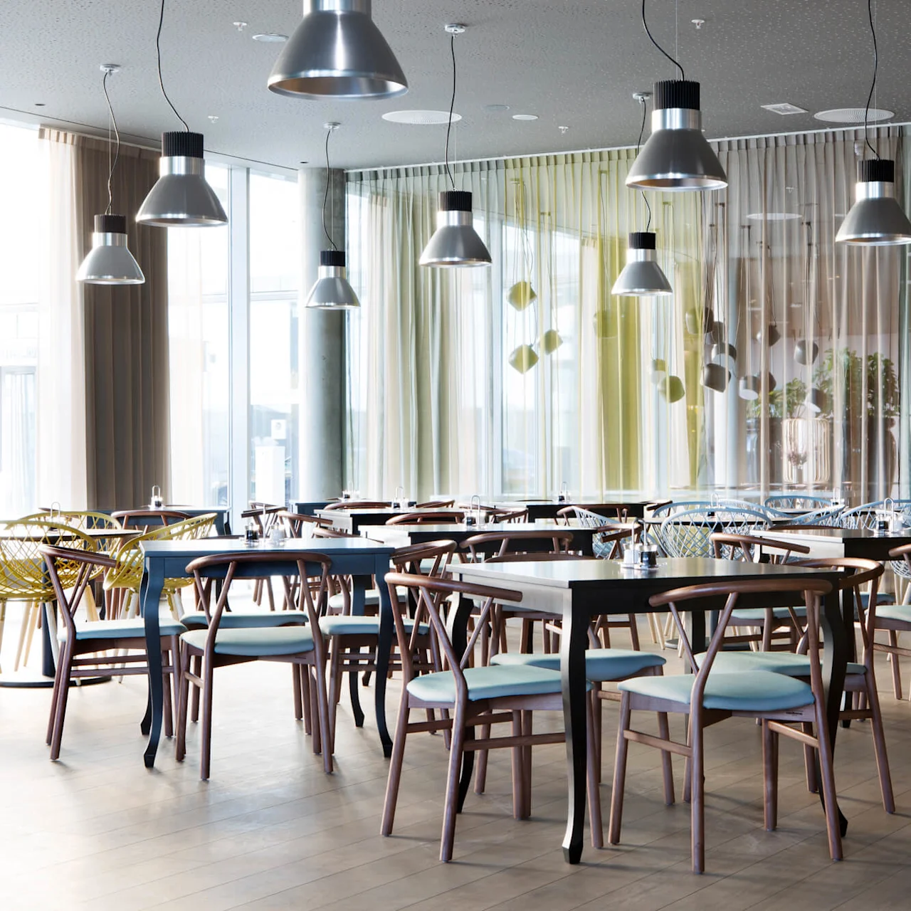 Tables and chairs arranged neatly in a modern, spacious dining area with large windows and hanging lamps, creating a well-lit, airy atmosphere.
