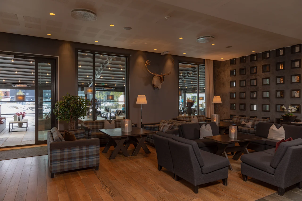 Koselig lounge med rutete lenestoler og sofaer arrangert rundt trebord, opplyst av varme gulvlamper. Veggene har firkantede dekorative aksenter og hjortegevir. Store vinduer viser en utendørs sitteplass.