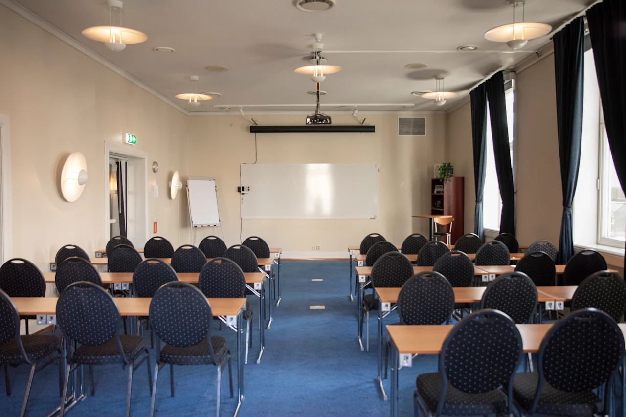 Et rummeligt konferencelokale med rækker af borde og stole, et projektorlærred og en whiteboardtavle, klar til en præsentation.