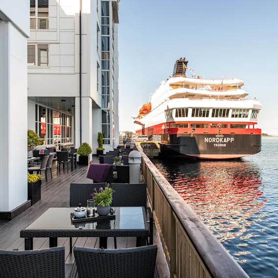 Et stort skip merket "NORDKAPP TROMSØ" er fortøyd ved en moderne bygning ved vannkanten. Utendørs sitteplasser med bord og planter langs en tredekke.