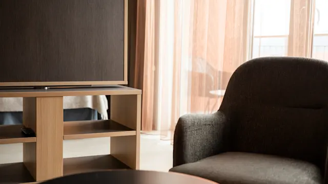 En mørk polstret lenestol står ved siden av et svart rundt bord i en stue. Et TV-stativ i tre med hyller er synlig, og tynne gardiner slipper inn mykt lys.