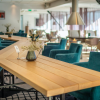 Wooden tables and teal chairs in a bright, modern dining area with large windows and soft lighting.