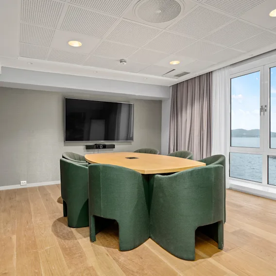 Et rundt bord med grønne stoler er plassert nær store vinduer i et moderne møterom. Rommet har en veggmontert TV og lys tregulv.