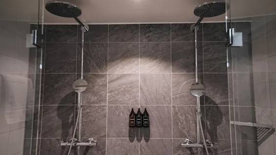 Et moderne dusjområde med to dusjhoder, mørke flislagte vegger og tre flasker toalettartikler.