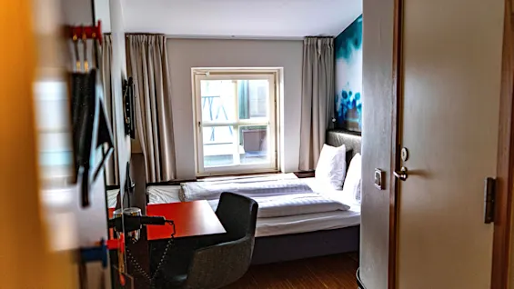 Ett mysigt hotellrum med en säng, ett litet skrivbord och ett fönster. Rummet känns kompakt och funktionellt.