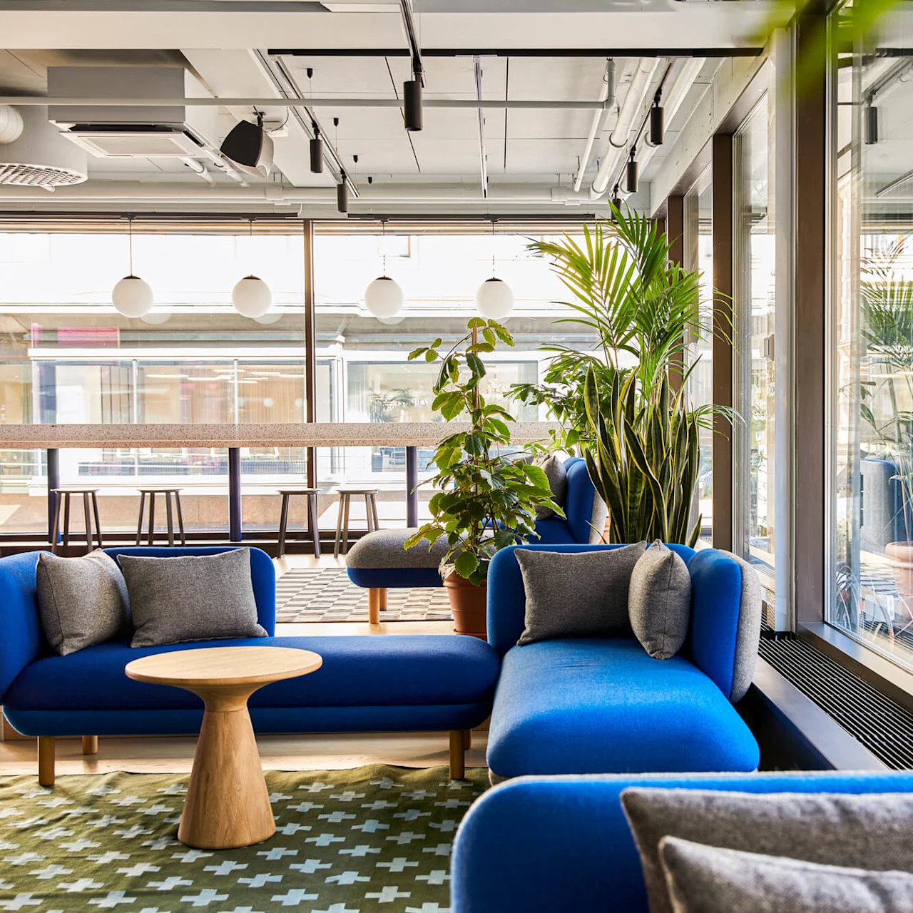 Blå sofaer med grå puder vender mod et lille træbord, omgivet af indendørs planter og store vinduer i et moderne loungeområde med lys ovenover.