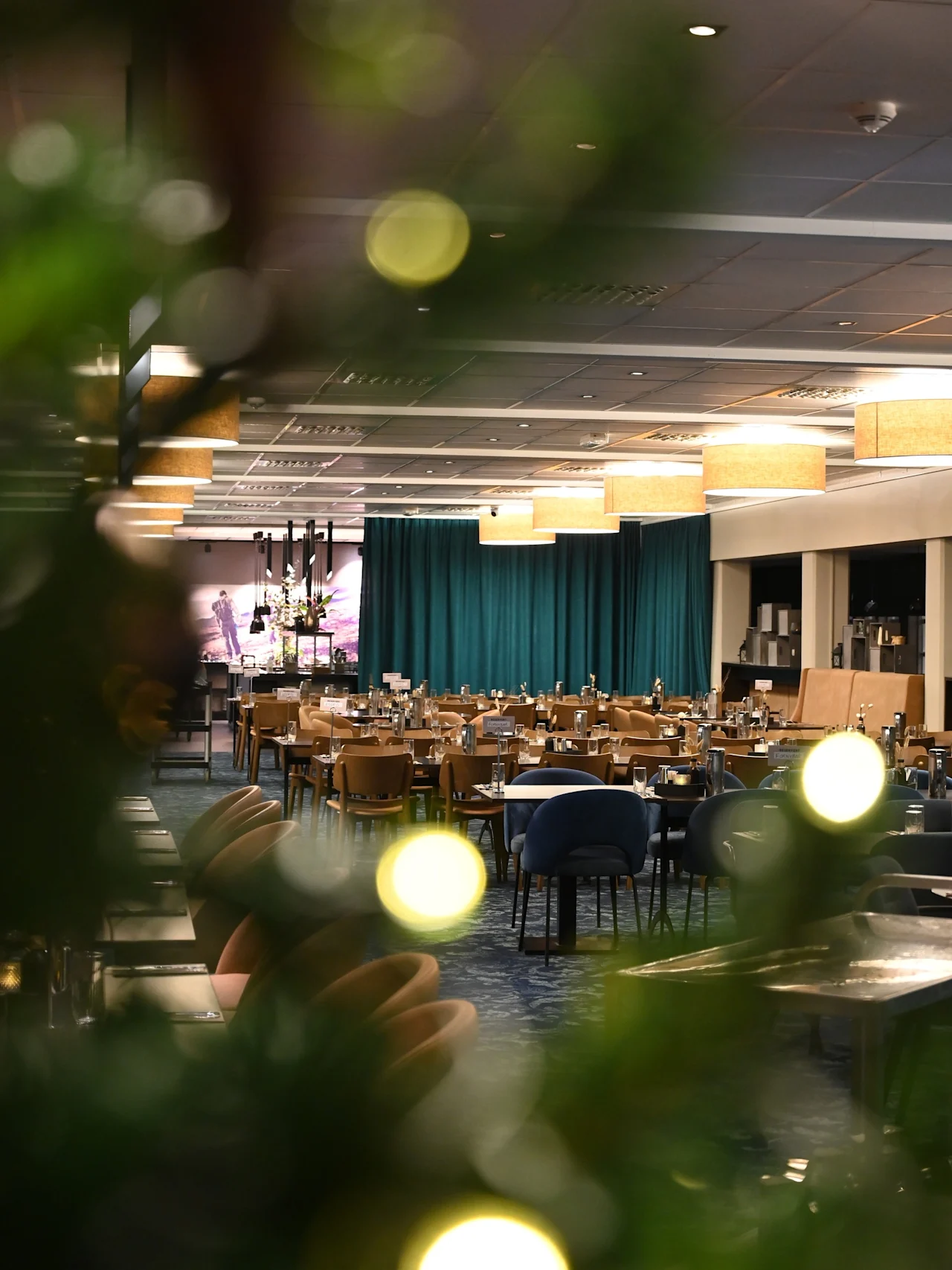 En spisesal fylt med pent arrangerte bord og stoler, under myk belysning. Grønt løvverk med uskarpe festlys rammer inn forgrunnen, mens turkise gardiner dekker bakveggen.