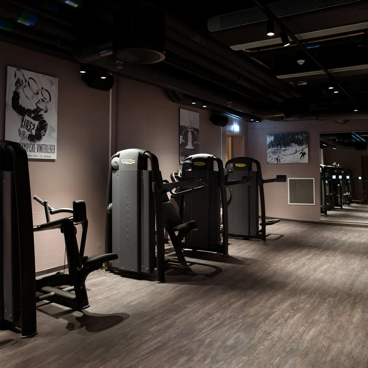 Fitness machines stand in a dimly lit gym with wooden floors and mirrored walls; posters of Olympic athletes decorate the walls. Text: "OLYMPISKE VINTERLEKER."