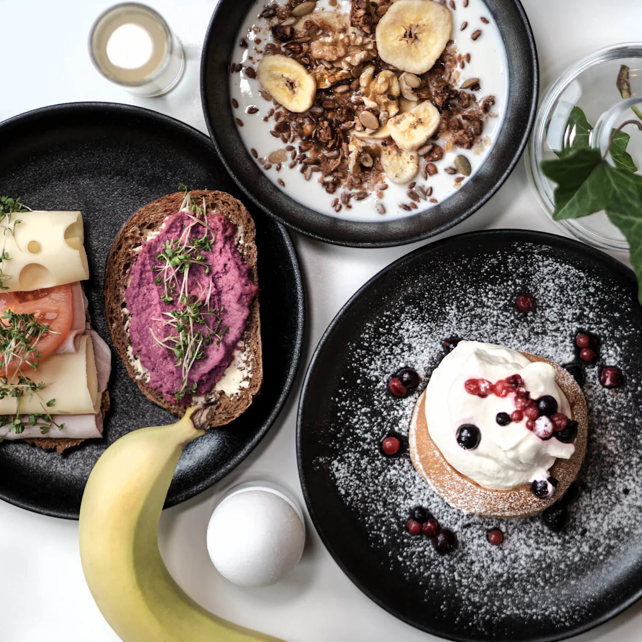 Pannkakor med grädde och bär serveras på en svart tallrik, omgivna av en banan, smörgåsar, granola, juice och en krukväxt på ett vitt bord.