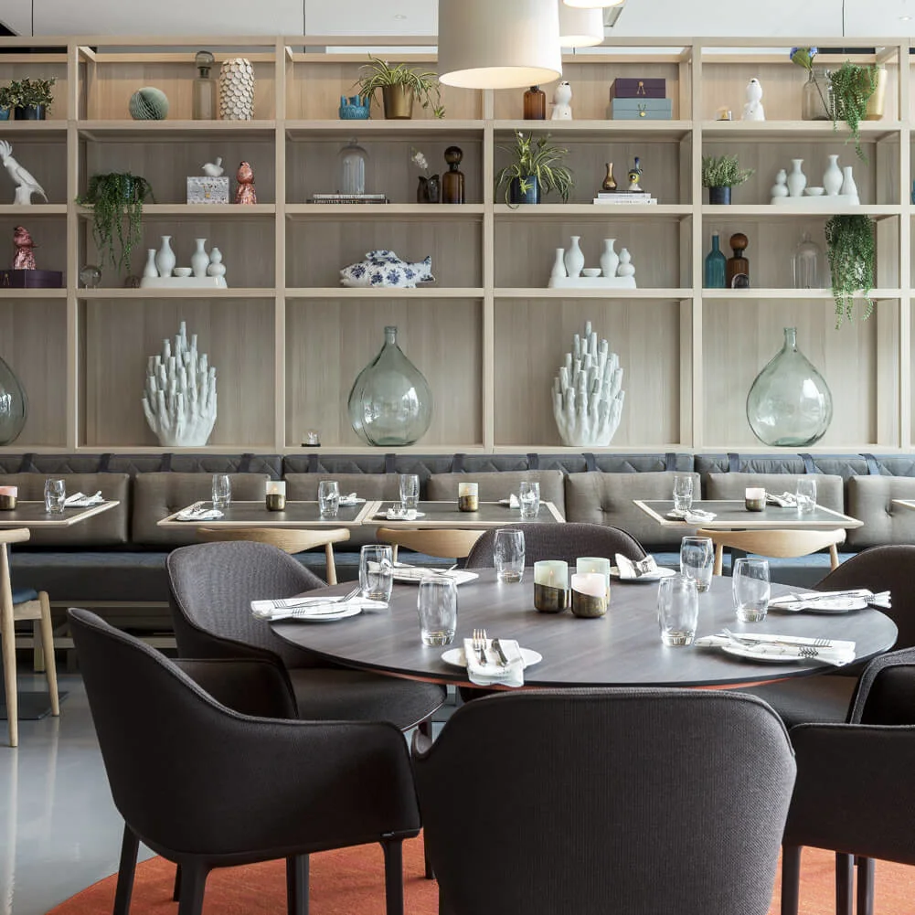 A round dining table set with glasses and napkins sits beneath pendant lights. In the background, a shelving unit displays plants and decorative items.