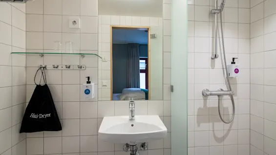 Et badeværelse med hvide flisebelagte vægge. Vasken og spejlet er centralt, under en væglampe. En hårtørrerpose, hylde med glas ovenover, brusehoved og sæbedispensere er synlige.