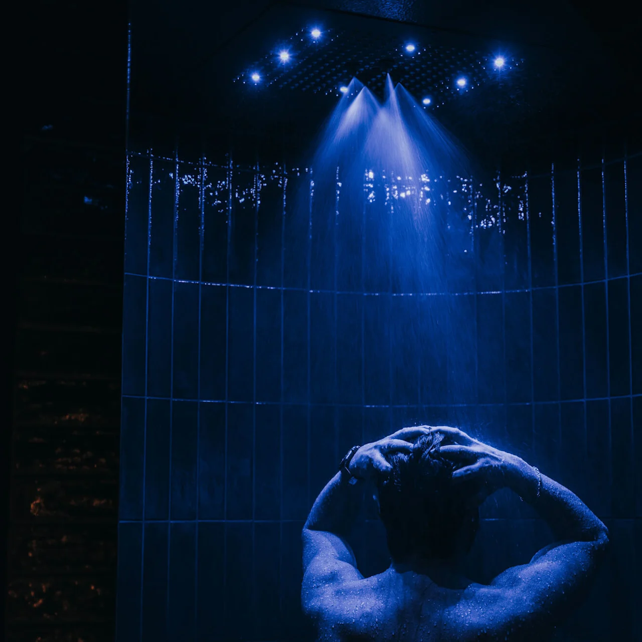 Person showering under blue LED-lit water streams, hands on head, standing in a dark tiled bathroom.