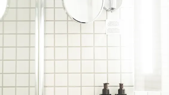 Väggmonterad rund spegel reflekterar ljus, åtföljs av två mörka flaskor märkta "BODY LOTION" och "BODY WASH" på en kaklad badrumsvägg.