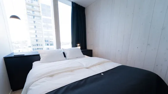 A neatly made bed with white linens and a dark bedspread sits in a minimalist room with light wood panel walls, next to a large window showing an outside building.