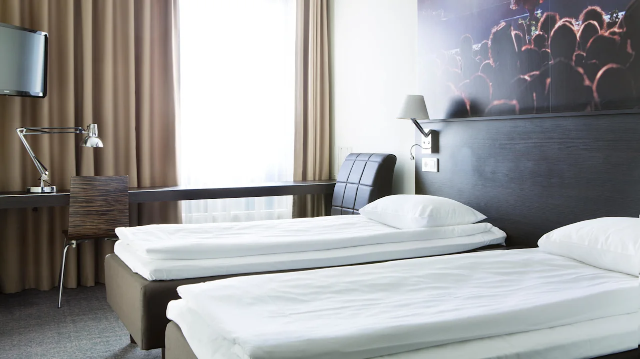 Two neatly made single beds with white linens stand in a modern hotel room. A desk with a lamp and chair sits beside a window with beige curtains.