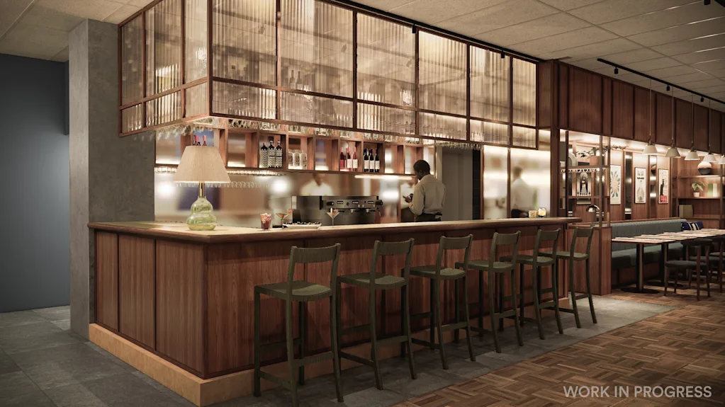 A person works behind a well-stocked bar with stools and a lamp. Tables and chairs are visible in the background. 