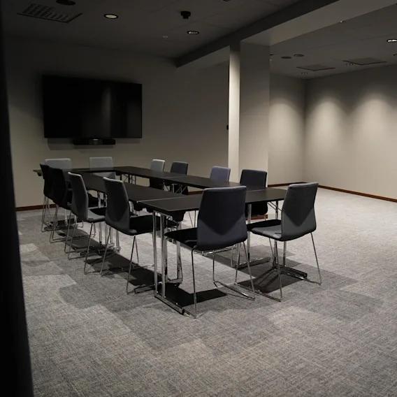 En rektangulär konferensbord, omgivet av mörka stolar, står i ett svagt upplyst, rymligt rum med heltäckningsmatta. En stor platt-TV är monterad på väggen.