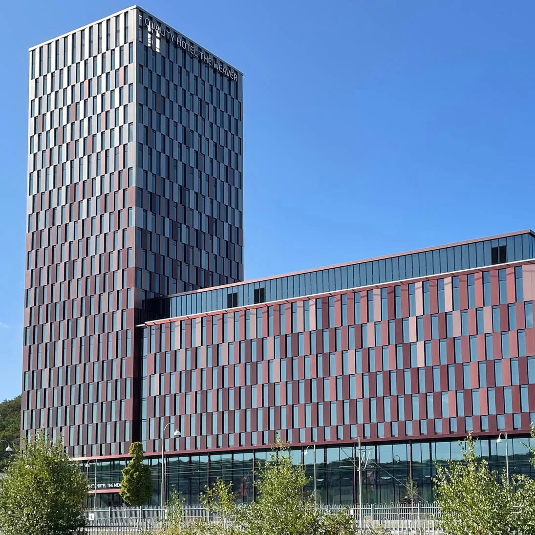 Ett modernt höghus visar "QUALITY HOTEL THE WEAVER" på fasaden, omgivet av träd under en klarblå himmel.