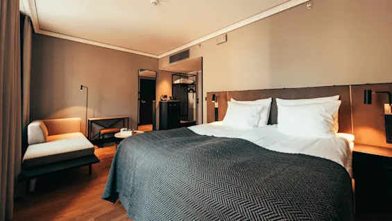 En pent, oppredd seng med hvite puter og et mørkt teppe står i et moderne hotellrom med en sjeselong, speil og et lite bord mot nøytraltfargede vegger.
